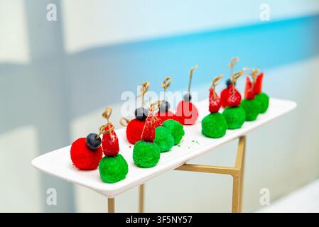 Hellrote und grüne Vorspeisenbällchen sind kunstvoll auf einem rechteckigen weißen Teller angeordnet, garniert mit Blaubeeren und Erdbeeren auf kleinen Spießen Stockfoto