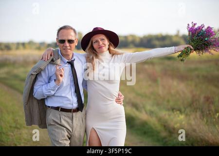 Ein Mann und eine Frau posieren zusammen auf einem grünen Feld an einem sonnigen Nachmittag. Die Frau hält einen Blumenstrauß und lächelt strahlend, während der Mann steht con Stockfoto