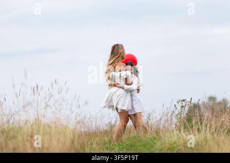 Eine Mutter umarmt ihre Tochter auf einer Wiese, beide lächeln, während sie einen leuchtend roten Ballon halten. Die Szene fängt einen warmen und fröhlichen Moment unter einem clou Stockfoto
