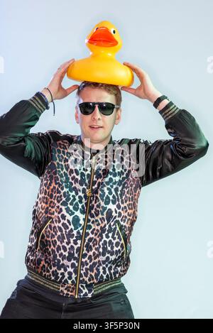Joe Lycett steht vor einem hellen Hintergrund und hält eine gelbe Gummiente auf dem Kopf. Er trägt eine Sonnenbrille und eine schwarze Jacke mit einem ausgeprägten Muster. Stockfoto