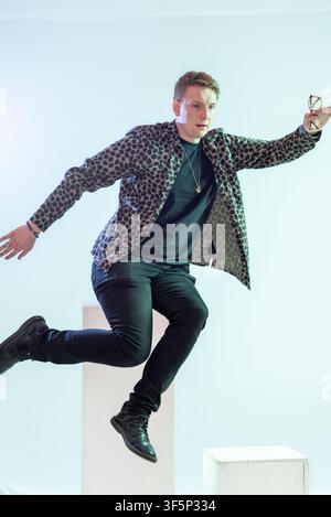 Joe Lycett wird in der Luft festgehalten, während er in einem Studio springt. Er hält eine Brille und trägt eine gemusterte Jacke über einem schwarzen Hemd. Der Hintergrund ist Stockfoto