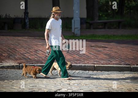 Kiew, Ukraine - 10. Jul 2026: Barfüßiger Mann mit Strohhut führt zwei Hunde an Leinen über den historischen Ziegelsteinpflasterweg des Mikhaylivska-Platzes Stockfoto