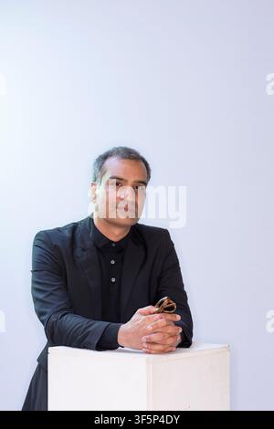 Nadeem Aslam steht mit verschränkten Armen auf einem weißen Podest in einer minimalistischen Umgebung. Er hält eine Brille und wirkt nachdenklich, während er schaut Stockfoto