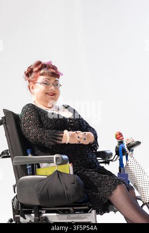 Penny Pepper sitzt im Rollstuhl, lächelt und trägt ein schwarzes Kleid. Sie befindet sich in einer einfachen Umgebung mit hellem Hintergrund. Sie strahlt Selbstvertrauen aus und ein Stockfoto