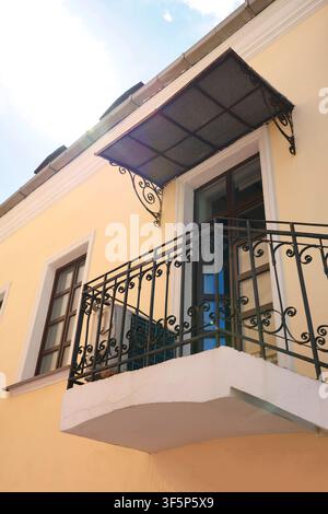 Schmiedeeiserner Balkon mit Schutzdach an gelber Gebäudefassade. Vintage-Metallgeländerrahmen und Polycarbonat-Markise über Glastür Stockfoto