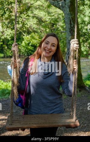 Junge Frau lächelt, während sie auf einem Seilschaukel in einem grünen Wald an einem Fluss sitzt Stockfoto