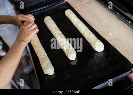 Professioneller Bäcker bereitet frische Teigbrote auf einem schwarzen Metalltablett in einer gewerblichen Kücheneinrichtung vor, während er rohe lange Brotstücke für den o formt Stockfoto