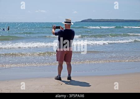 Rückansicht älterer Mann in Shorts und Sonnenhut, der am Strand von Marloes Sands in Pembrokeshire, Wales, UK, Wellen im Meer filmt und fotografiert  KATHY DEWITT Stockfoto
