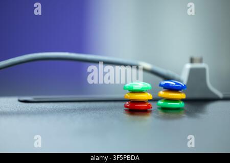 Bunte Stapel runder Plastikknöpfe, die auf einer glatten dunklen Oberfläche in der Nähe eines elektronischen Kabels mit verschwommenem Hintergrund und einer Farbverlaufswand im Inneren angeordnet sind Stockfoto