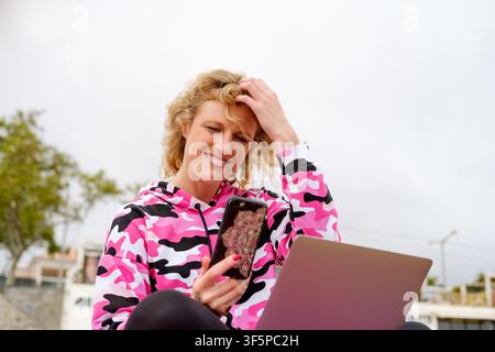 Frau lächelt, interagiert glücklich mit ihrem Smartphone und Laptop, während sie im Freien arbeitet Stockfoto