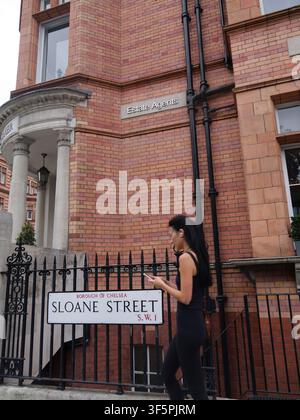 Eine modisch gekleidete junge Fußgängerin geht an einem traditionellen, wandmontierten Straßenschild für Sloane Street an einer Gebäudefassade im wohlhabenden Viertel Chelsea in London, UK, vorbei Stockfoto