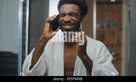 Glücklicher afroamerikanischer Mann, optimistisch, der am Handy telefoniert und seinen Spiegelreflex im Badezimmer zu Hause betrachtet, ein lächelnder Typ, der ein Smartphone-Gespräch führt und sein Gesicht berührt Stockfoto