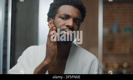 Portrait eines selbstbewussten jungen afroamerikanischen Mannes, der sanft seine Gesichtshaut und seinen Bart streichelt, in den Spiegelblick schaut und im Badezimmer einen Bademantel trägt Stockfoto