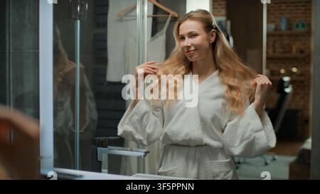 Glückliche schöne lächelnde kaukasische Frau mittleren Alters, die ihr langes blondes Haar berührt und sich im häuslichen Badezimmer auf ein Date vorbereitet. Modische attraktive Frau Stockfoto