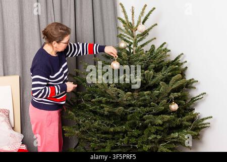 Eine Person fügt einem Weihnachtsbaum weiße Schmuckstücke hinzu. Der Baum steht in einem Wohnzimmer mit grauen Vorhängen. Die Aktivität findet während der Feiertage statt Stockfoto