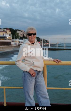 Schöne reife Frau in Sonnenbrille und lässigem weißen Hoodie, die sich an einem europäischen Küstenort am Abend an Metallgeländern lehnt. Stockfoto