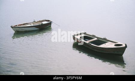 Zwei verwitterte Holzruderboote treiben auf ruhigem, nebeligem Wasser und schaffen eine stille, nostalgische Meereslandschaft Stockfoto