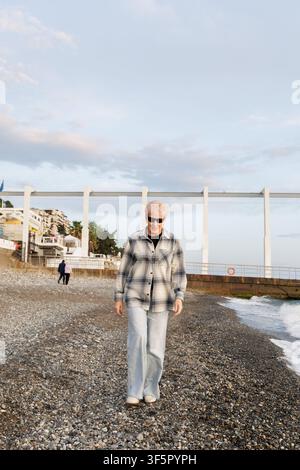Ein ungeposester Mittelformat-Bild einer nachdenklichen Seniorin in einer karierten Jacke, die während eines entspannenden Urlaubs an einer Küste spaziert. Stockfoto