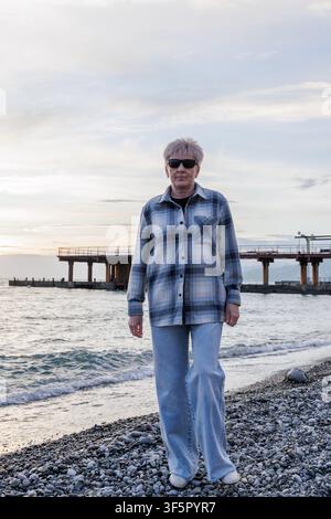 Ein ungeposester Mittelformat-Bild einer nachdenklichen Seniorin in einer karierten Jacke, die während eines entspannenden Urlaubs an einer Küste spaziert. Stockfoto