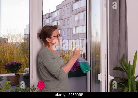 Eine Person wäscht Fenster in einer Wohnung auf einem mit Blumen gefüllten Balkon. Diese Aktivität findet im Herbst statt, wenn der Winter naht, um sich auf die kälteren Monate vorzubereiten Stockfoto