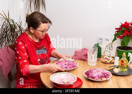 Frau bereitet Olivier-Salat für Weihnachten vor. Sie arrangiert sorgfältig Schichten von gekochtem Gemüse und fügt Mayonnaise in einer gemütlichen Kücheneinrichtung hinzu, umgeben von Pflanzen Stockfoto