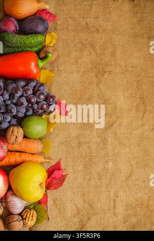 Flat Lay mit verschiedenen frischen Herbstgemüsen, Früchten und Nüssen, die entlang der linken Seite eines rustikalen Sackleinenhintergrunds angeordnet sind, mit großzügigem vertikalem Platz für Text Stockfoto