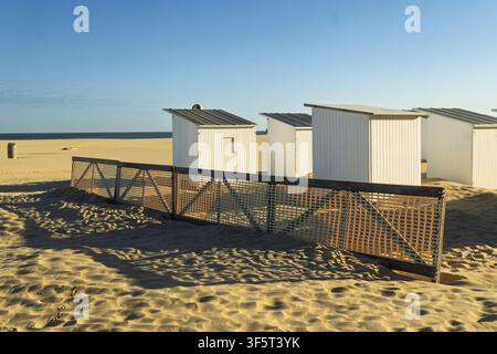 Weiße Hütten mit grauen Dächern stehen auf dem hellen Ostender Sand, mit Netzen, die den Sand zurückhalten, zum weiten Strand und der ruhigen See unter einem klaren blauen Himmel gerichtet. Stockfoto