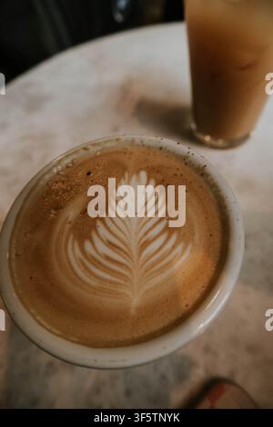 Nahaufnahme eines Lattes mit Rosetten-Schaumkunst auf einem Marmor-Café-Tisch, mit einem weich unscharf im Hintergrund stehenden Eiskaffee Stockfoto