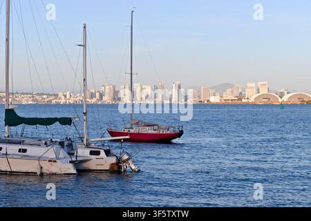 Mehrere Segelboote ruhen auf blauem Wasser in der San Diego Bay, während sich die Innenstadt-Skyline und die Ufergebäude unter einem klaren Nachthimmel erheben. Stockfoto