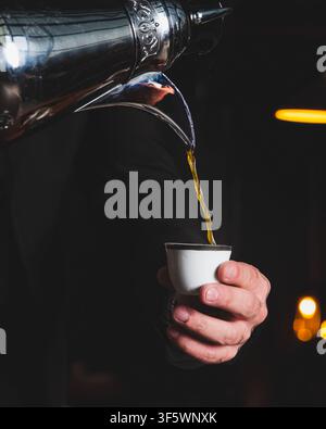 Goldener arabischer Kaffee wird aus einem verzierten silbernen Topf in eine traditionelle finjan-Tasse ohne Henkel gegossen, die ein Mann in der Hand hält, vor einem dunklen Hintergrund. Stockfoto