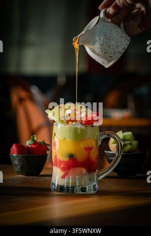 Honig wird über einen geschichteten gemischten Obstcocktail gegossen, der in einer Glas Tasse mit Ananas, Erdbeere und Sahne gekrönt ist, mit Schüsseln mit frischen Früchten. Stockfoto