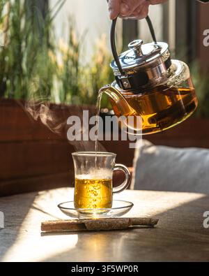 Heißer Kräuter-Zimttee, der aus einer gläsernen Teekanne in ein traditionelles Istikan-Glas gegossen wird, mit aufsteigendem Dampf, von goldenem Sonnenlicht hinterleuchtet auf einem Café-Tisch. Stockfoto