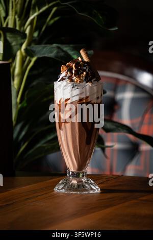 Schokoladenmilchshake mit Schlagsahne, Schokoladensoße, gehackten Nüssen und einer Waffelrolle, serviert in einem hohen Fußglas auf einem Holztisch. Stockfoto