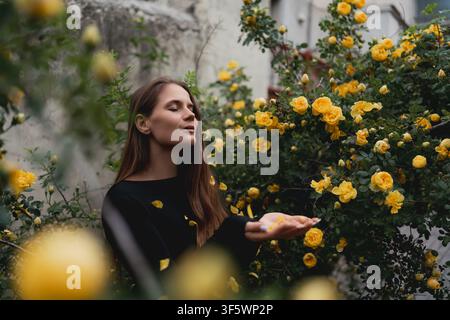 Junge Frau genießt fallende Blütenblätter von gelben Rosen, während sie in einem üppigen Sommergarten steht Stockfoto