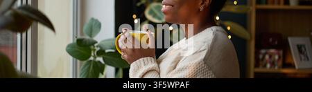 Afroamerikanische junge Erwachsene hält eine gelbe Tasse und schaut aus dem Fenster, lächelt während der morgendlichen Kaffeepause. Bild repräsentiert psychologisches Wohlbefinden und Selbstfürsorge-Marketingkampagnen Stockfoto
