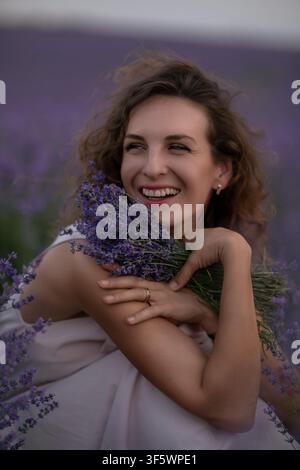Fröhliche junge Frau hält einen duftenden Lavandula-Strauß, während sie während eines malerischen Sommerabends in einem lila Feld sitzt Stockfoto