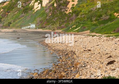 Felsige Küste und Küstenklippen erstrecken sich entlang von Rat Beach, Kalifornien, während zwei Personen in der Nähe von sanften Wellen und grünen Hügeln spazieren gehen. Stockfoto
