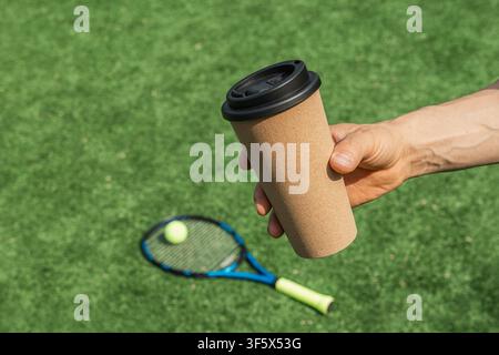 Hand, die einen To-go-Kaffeebecher hält, nach dem Tennistraining mit Schläger und Ball auf dem Außenplatz. Aktiver Lebensstil, Sportpause und gesunde Erholung mit Stockfoto