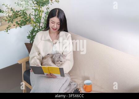 Asiatische Frau liest ein Buch, während sie mit einer Katze auf dem Sofa zu Hause entspannt Stockfoto