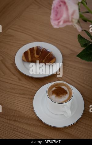 Cappuccino und frisches Croissant auf einem Holztisch mit weichen rosa Rosen, gemütliche Frühstücksszene in elegantem natürlichem Morgenlicht. Stockfoto