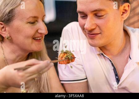 Frau füttert lächelndem Mann Sushi in einer gemütlichen Restaurantatmosphäre. Japanisches Sushi-Konzept für ein Pärchen beim Abendessen und romantisches asiatisches Esserlebnis. Stockfoto