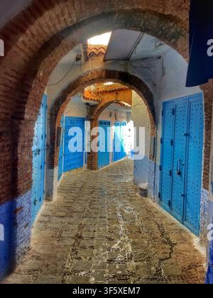 Schmale Kopfsteinpflastergasse mit blauen und ockergelben Wänden und einem gewölbten Tor in Chefchaouen, Marokko. 4. August 2025. Stockfoto
