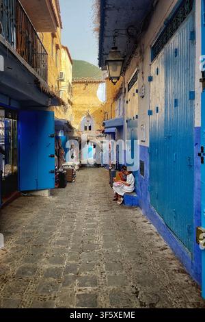 Schmale Kopfsteinpflastergasse mit blauen und ockergelben Wänden und einem gewölbten Tor in Chefchaouen, Marokko. 4. August 2025. Stockfoto