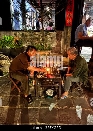 Zwei ältere Männer spielen chinesisches Schach (Xiangqi) an einem kleinen Holztisch entlang einer laternenbeleuchteten Straße in Hoi An, Vietnam, am 1. April 2026. Stockfoto