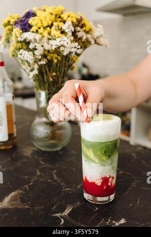 Hand, die einen gestreiften Papierstrohhalm in einen frisch zubereiteten geschichteten Erdbeer-Matcha-Latte legt. Erfrischendes Eiskaffeegetränk zum Servieren bereit. Stockfoto