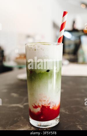Schichtweiser Erdbeer-Matcha-Latte mit cremigem Schaum gekrönt und mit einem gestreiften Papierstrohhalm serviert. Erfrischendes Spezialkaffegetränk im Nahaufnahme Stockfoto