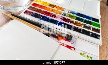Hohe Blickwinkelaufnahme von Wasserfarben-Materialien mit einem farbenfrohen Farbsatz, Pinseln und einem leeren Notizbuch auf einem Holztisch. Konzept der Kreativität, Feins Stockfoto