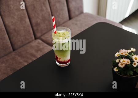 Schichtweiser Erdbeer-Matcha-Latte serviert über Eis mit cremigem Schaum und einem gestreiften Papierstrohhalm. Erfrischendes Spezialgetränk, präsentiert in einem modernen Café Stockfoto
