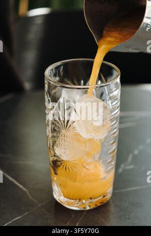 Frischer Orangensaft wird über Eiswürfel in ein dekoratives Glas gegossen. Erfrischendes Zitrusgetränk, zubereitet in einem modernen Café. Stockfoto