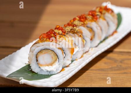 Exquisite Tempura-Garnelen-Sushi-Rollen, belegt mit rotem Kaviar, angebratenem Fisch und Gewürzen, serviert auf einem weißen langen Teller mit einer Bambusblatt-Garnitur. Stockfoto